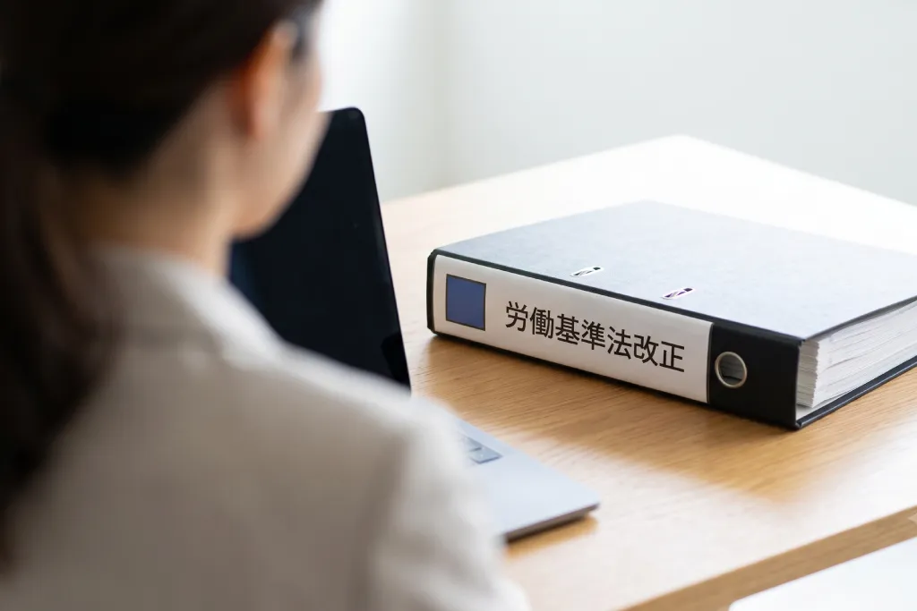 オフィスのデスクでパソコンに向かう女性の肩越しに写る、「労働基準法改正」と書かれた黒いビジネスバインダー